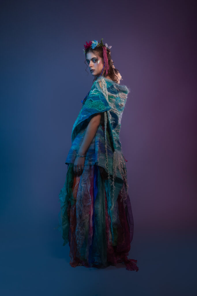 Performer wearing a layered, flowing costume in blue and green tones, photographed against a gradient studio background.